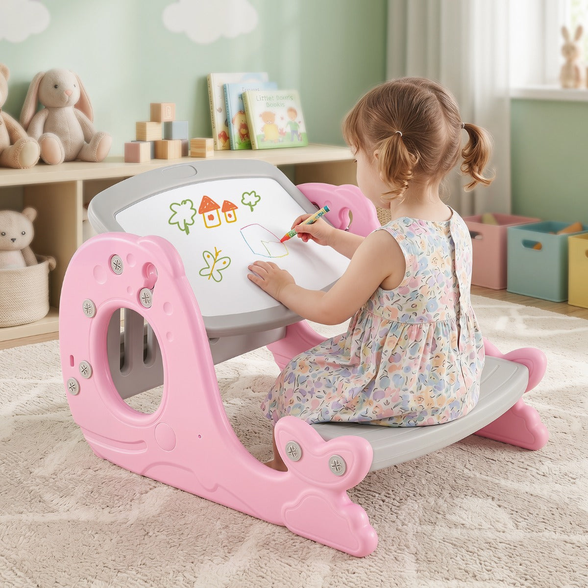 A young child sits on a pink and gray rocking writing desk in a playroom, drawing on the white writing surface with a colored pencil.