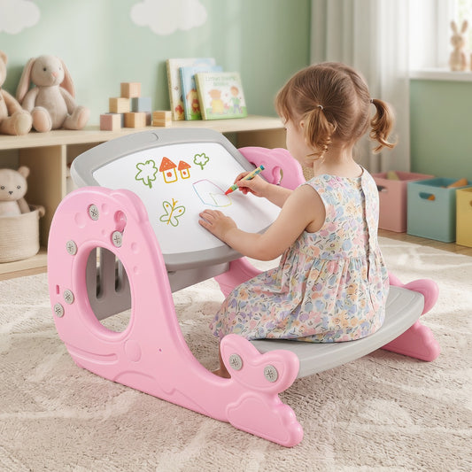 A young child sits on a pink and gray rocking writing desk in a playroom, drawing on the white writing surface with a colored pencil.